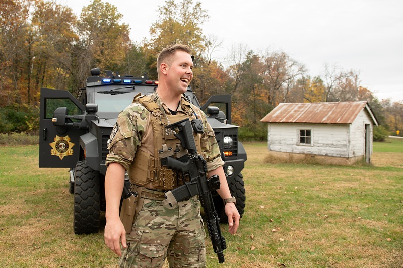 male officer in camouflage in front of armored vehicle.